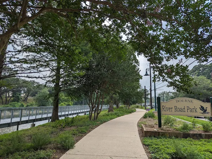 River Road Park Boerne Walking Trail and Bridge Walking path at River Road Park in Boerne, Texas, running alongside a river with a metal pedestrian bridge and park sign.